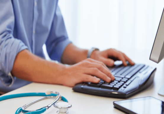 Close up image of male GP hands typing on a keyboard with stethoscope and phone next to him 